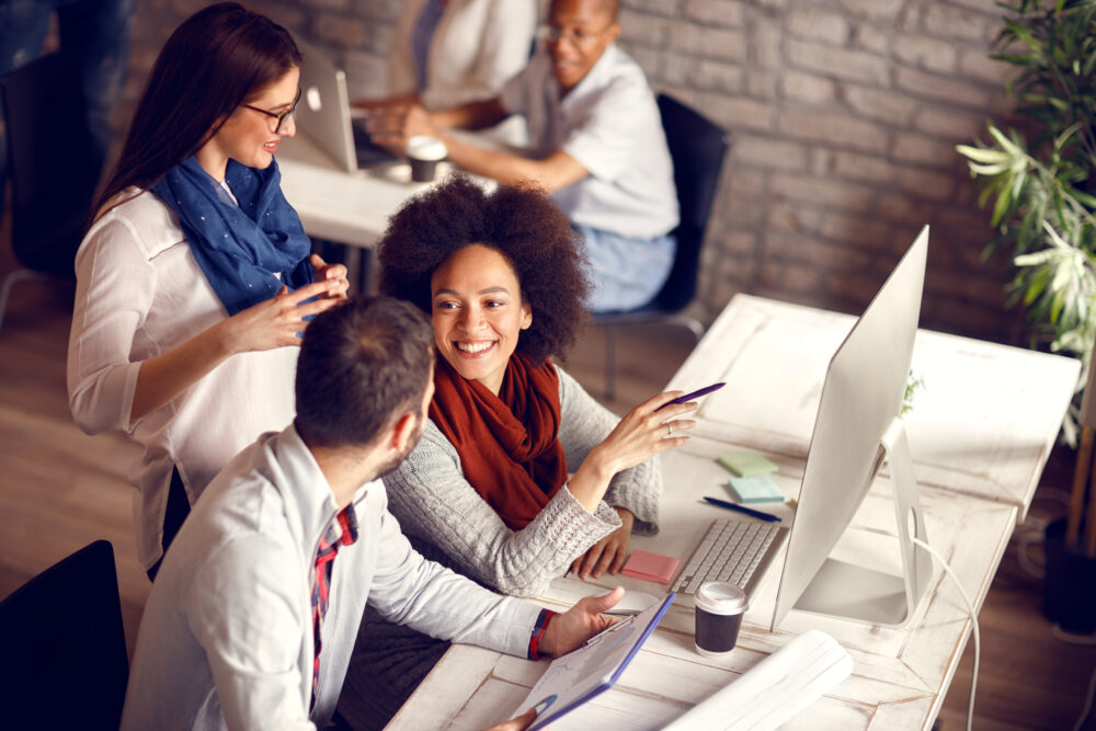 A group of three employees gather around a computer having a discussion. Another employee sits in the background.